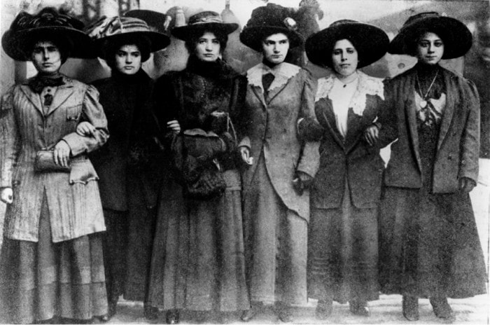 six women in nineteen hundreds dress link arms while marching.