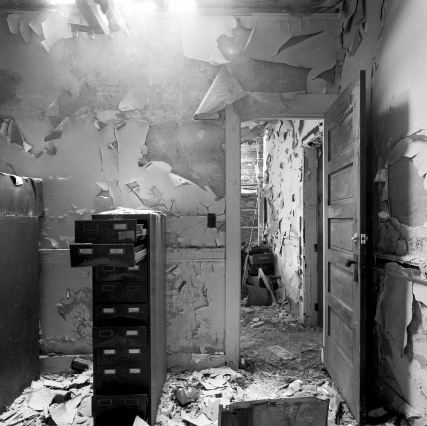 A photograph by Jeremy Blakeslee showing an abandoned police station in High Park, Detroit. A file cabinet stands open, papers litter the floor, paint peels from the walls.